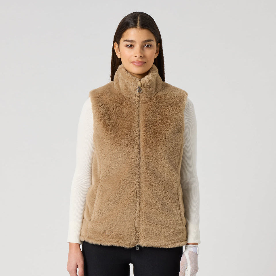 A woman modeling a tan sherpa fleece zip-up vest over a white long-sleeve top and black pants, standing against a light gray studio background.