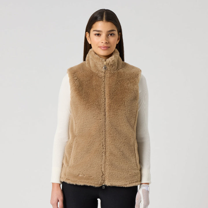 A woman modeling a tan sherpa fleece zip-up vest over a white long-sleeve top and black pants, standing against a light gray studio background.