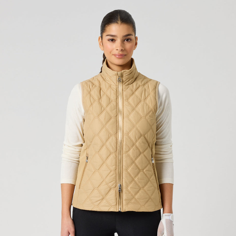 A woman modeling a quilted sleeveless vest in a light tan/beige color, worn over a white long-sleeve top and black pants, against a plain light gray studio background.