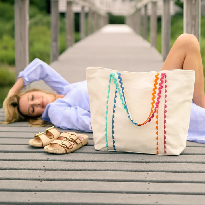 A woman lies on a wooden dock beside water, with a large cream canvas tote bag featuring colorful pom-pom handles and a pair of beige sandals in the foreground.